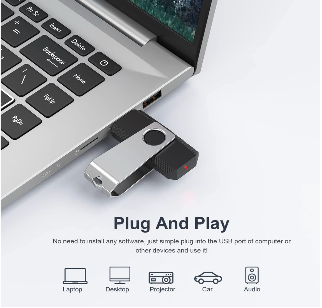 A silver and black USB flash drive plugged into a laptop USB port, with text below reading 'Plug And Play' and icons showing compatibility with laptop, desktop, projector, car, and audio devices.